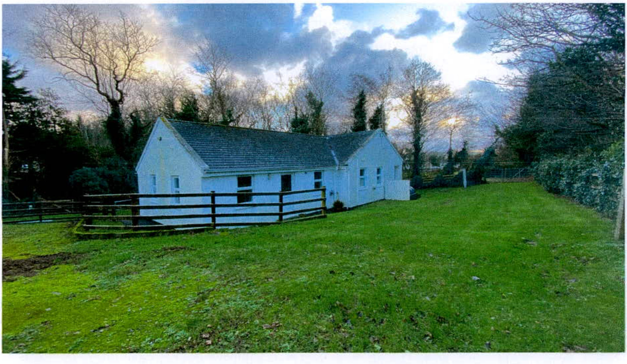 A photograph showing a white, single-story detached building with a pitched roof, situated in a grassy rural setting with a wooden fence and trees.