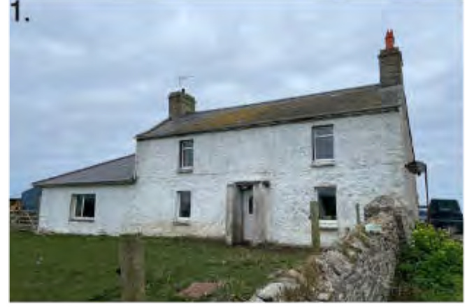 A photograph of an existing white two-story stone cottage with a single-story extension, situated in a rural setting with a stone wall in the foreground.