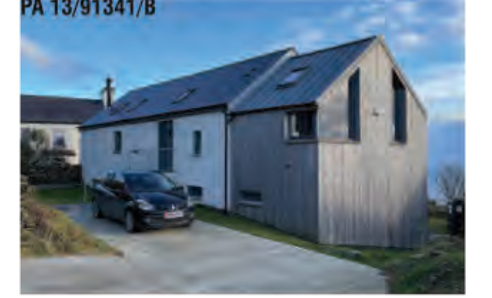 A photograph of a modern rural dwelling, appearing to be a barn conversion, featuring mixed cladding, roof-mounted solar panels, and a car parked in the driveway.