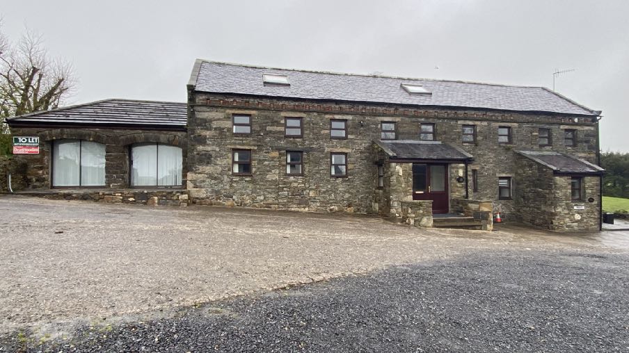A photograph of a stone building complex with a gravel forecourt, featuring a mix of traditional stone walls and modern extensions with large windows.