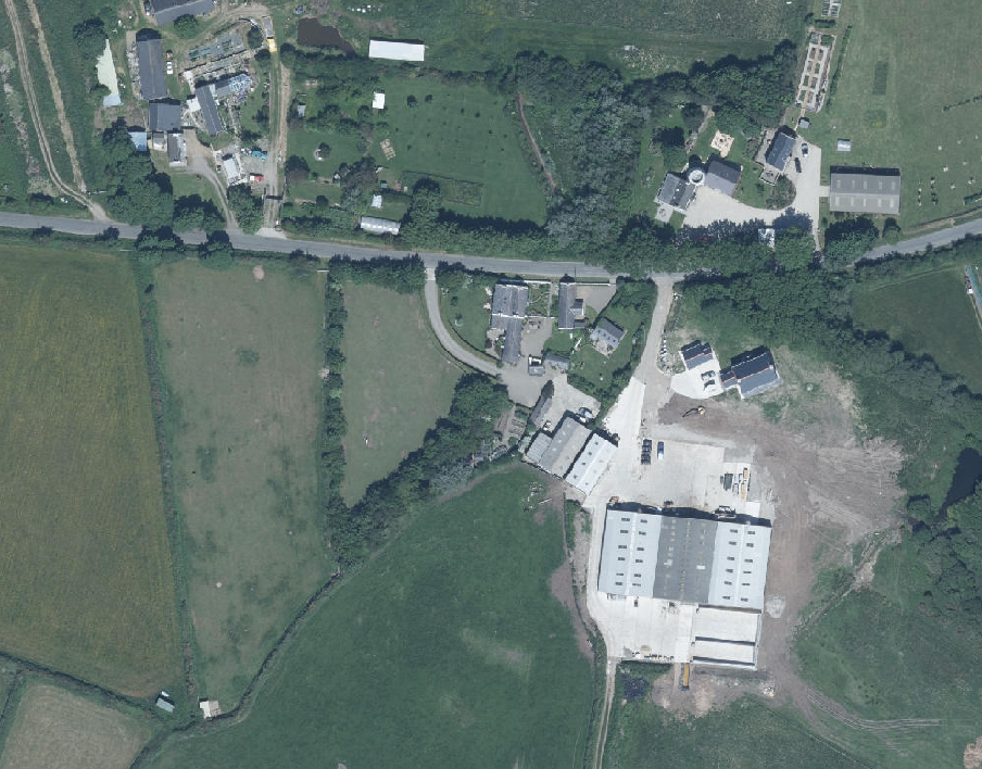 Aerial view of a rural site showing agricultural fields, a road, and existing buildings including large agricultural structures.