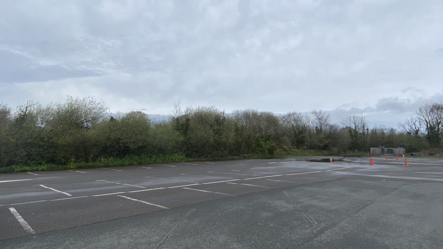 A photograph showing a paved parking area with white bay markings, bordered by a dense line of trees and bushes under an overcast sky.