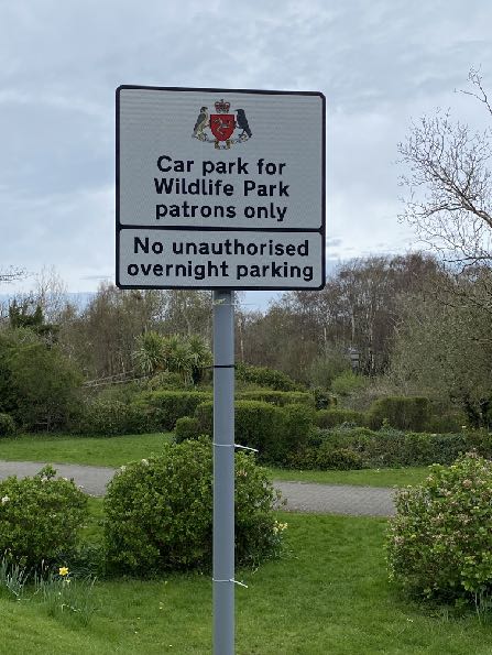 A photograph of a white signpost stating 'Car park for Wildlife Park patrons only' and 'No unauthorised overnight parking' set against a backdrop of greenery and trees.