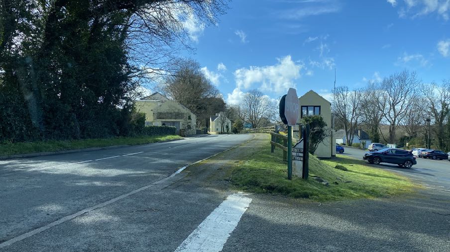 A street-level photograph showing a rural road junction with a stop sign, trees, and buildings in the background.