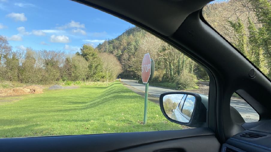A photograph taken from inside a vehicle showing a rural road junction with a stop sign and wooded hillside in the background.