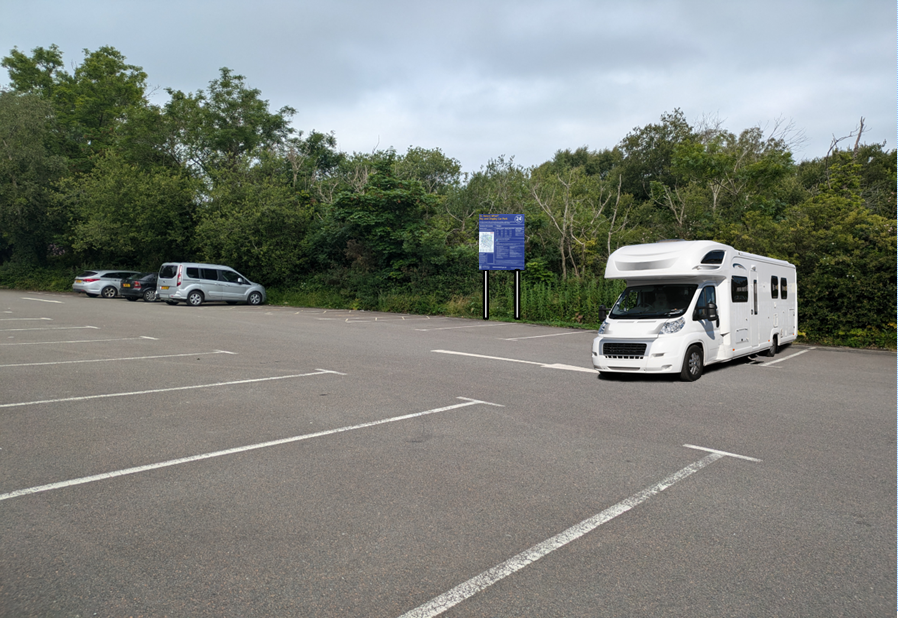 A photograph of a paved parking area with white bay markings, featuring a large white motorhome parked in the foreground and other vehicles in the background near a tree line.