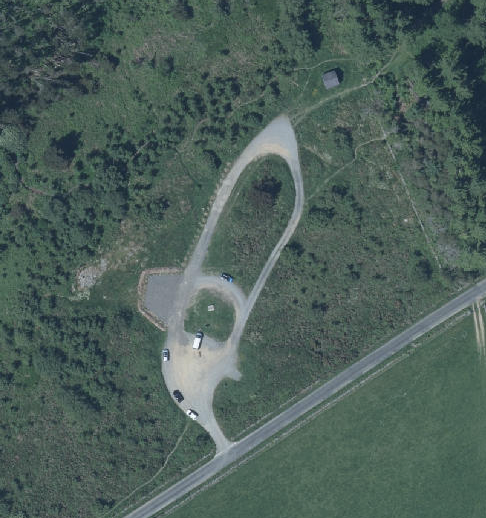 An aerial view of a rural property showing a looped driveway and parking area with several vehicles parked near a main road.