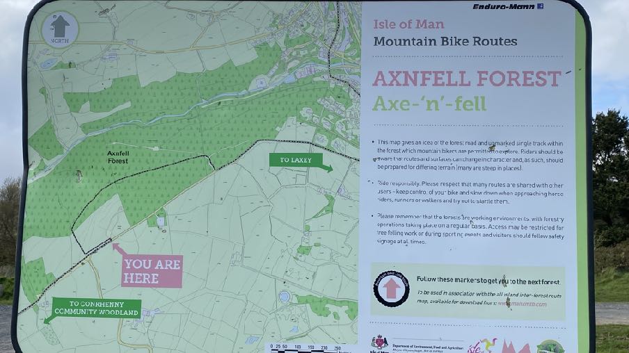 A photograph of an information sign displaying a map of the Axonfell Forest mountain bike routes with a 'You Are Here' marker.