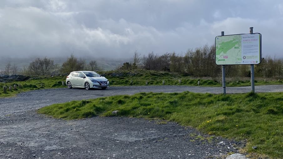 A photograph showing a gravel parking area with a white car and a large information sign board displaying a map.