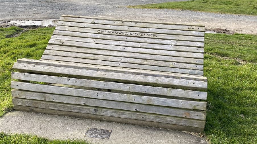 A photograph of a wooden bench with 'CELEBRATING OUR DARK SKIES' carved into the backrest, situated on a concrete pad next to grass and a gravel path.