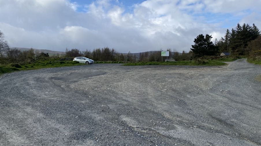 A wide-angle photograph of a gravel parking area or layby in a rural setting, featuring a white car parked on the left side with hills and trees in the background.