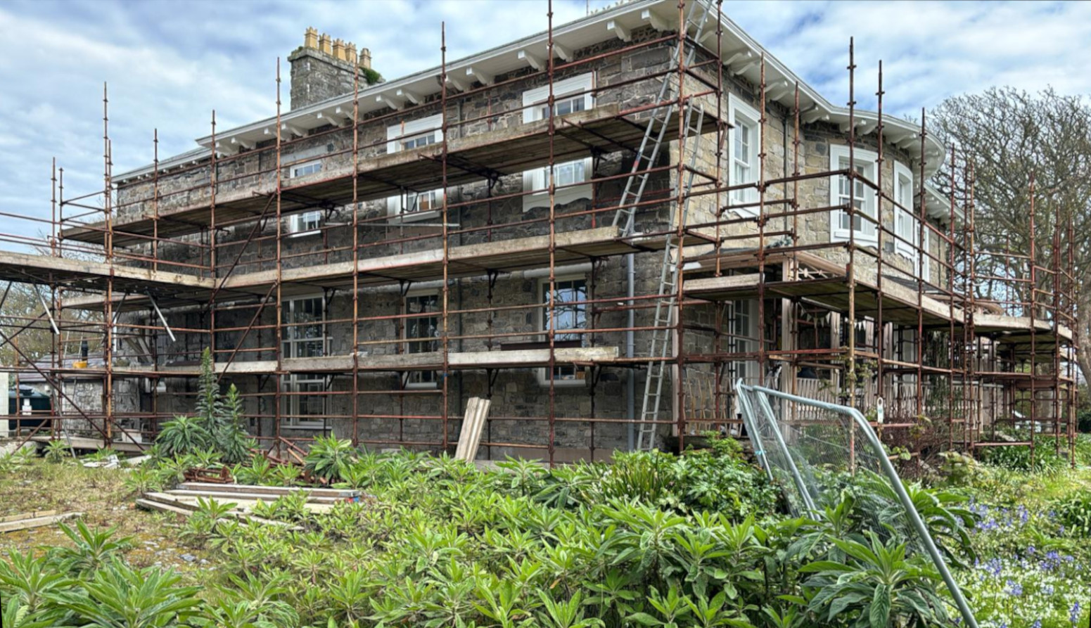 A large, multi-story stone house is fully covered in scaffolding, indicating significant renovation or construction work. The foreground features a garden with lush greenery and a metal frame structure.