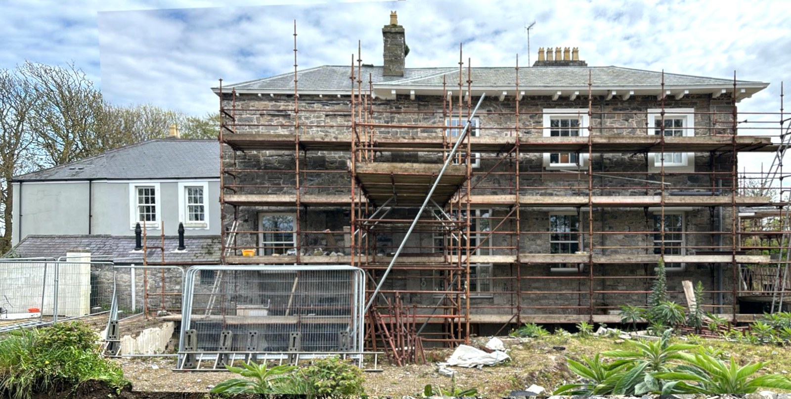 A photograph showing a large stone building covered in scaffolding with construction fencing and landscaping work visible in the foreground.