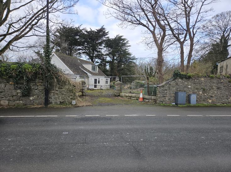 A street-level photograph showing the existing white detached bungalow behind stone boundary walls and a metal gate on Mill Road.