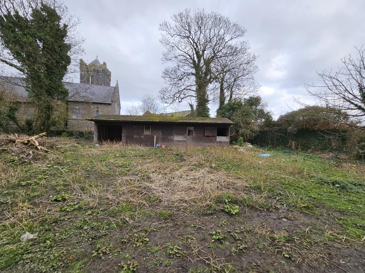 A photograph showing a grassy plot of land with a dilapidated wooden outbuilding or workshop, with a stone church visible in the background.
