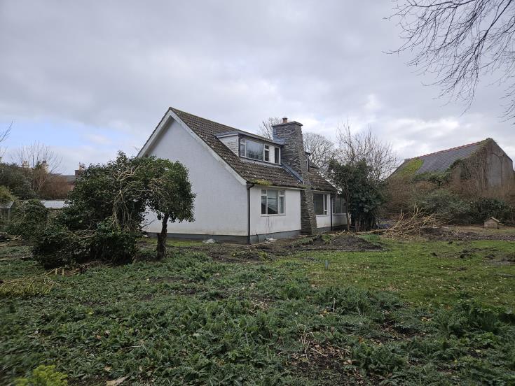 A photograph showing the existing white detached bungalow proposed for demolition, featuring a pitched roof, dormer window, and a garden area in the foreground.