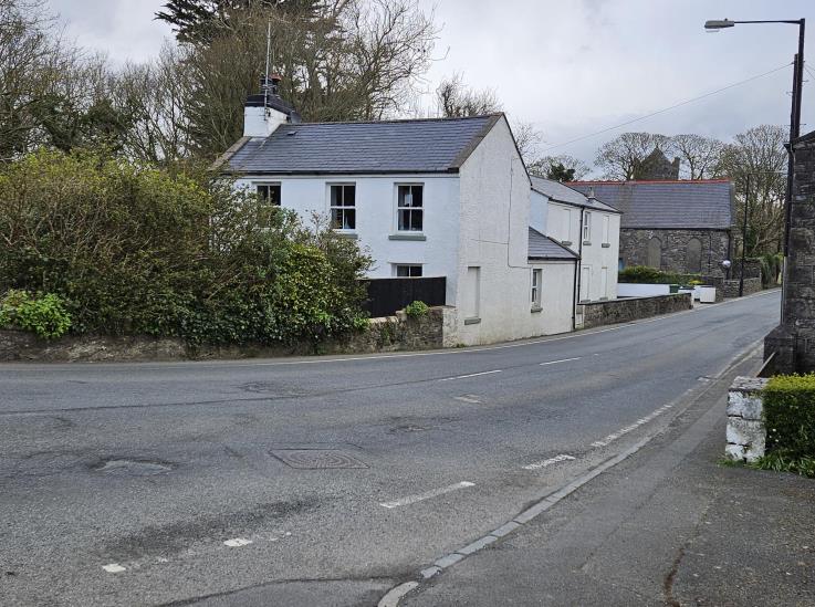 A street-level photograph showing an existing white two-story detached house on a corner plot with a slate roof, situated next to a stone building.