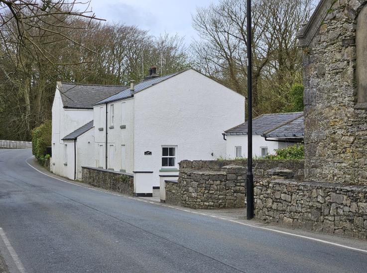 A street-level photograph showing a white residential building situated on a bend in a rural road, bordered by a stone wall and trees.