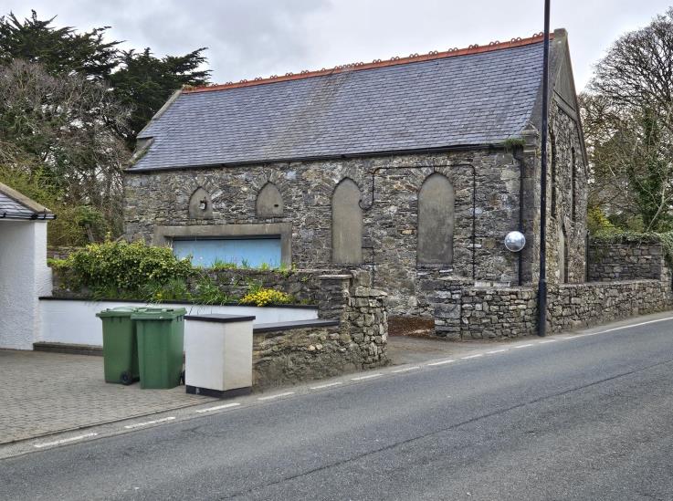 A street-level photograph of an existing stone building with arched windows, situated next to a road with green bins in the foreground.