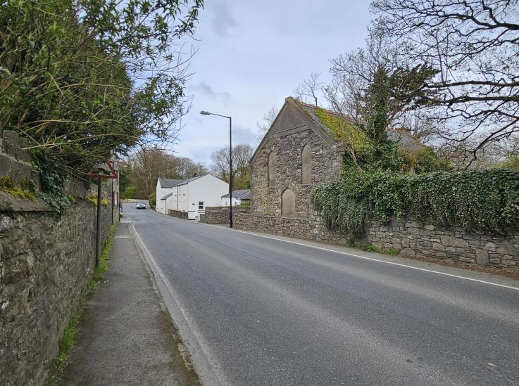 A street-level photograph showing a rural road scene with a stone building on the right and stone walls on the left, likely depicting the site of the proposed demolition.