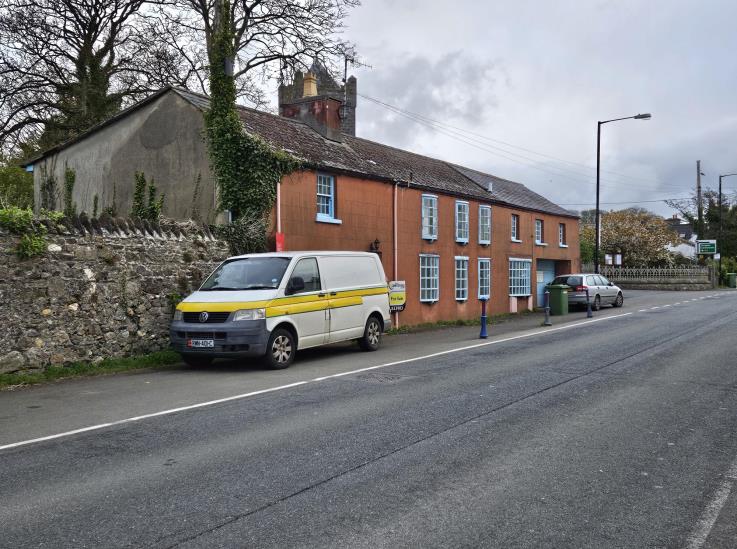 A street-level photograph showing a long red brick building and an adjacent grey structure on Mill Road, with a van parked in front and a stone wall in the foreground.
