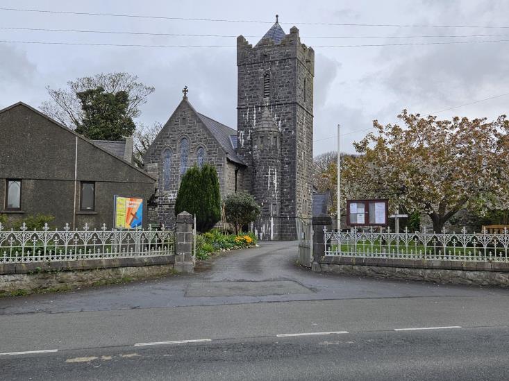 A street-level photograph showing a stone church with a tall tower and a gated entrance, alongside a grey building and white railings along the roadside.