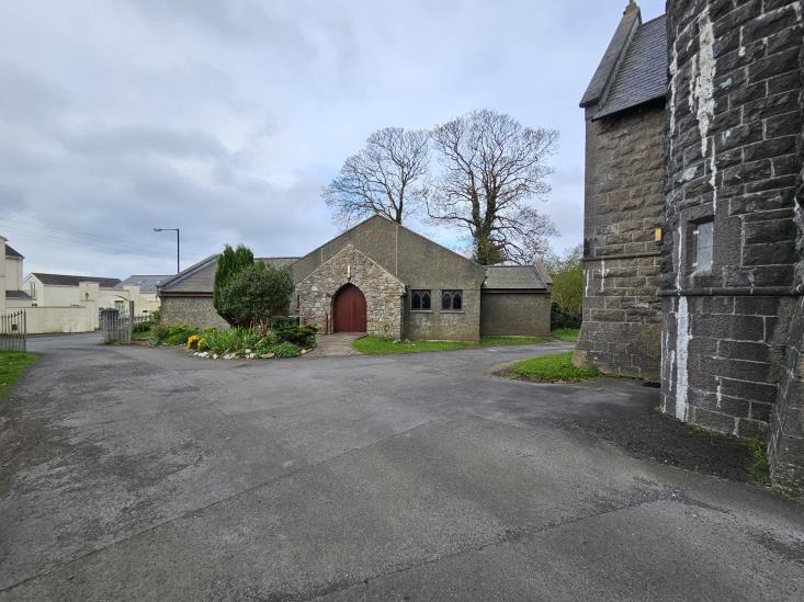 A photograph showing a stone building with a red arched door, likely a former chapel, situated next to a larger stone structure with a paved area in the foreground.