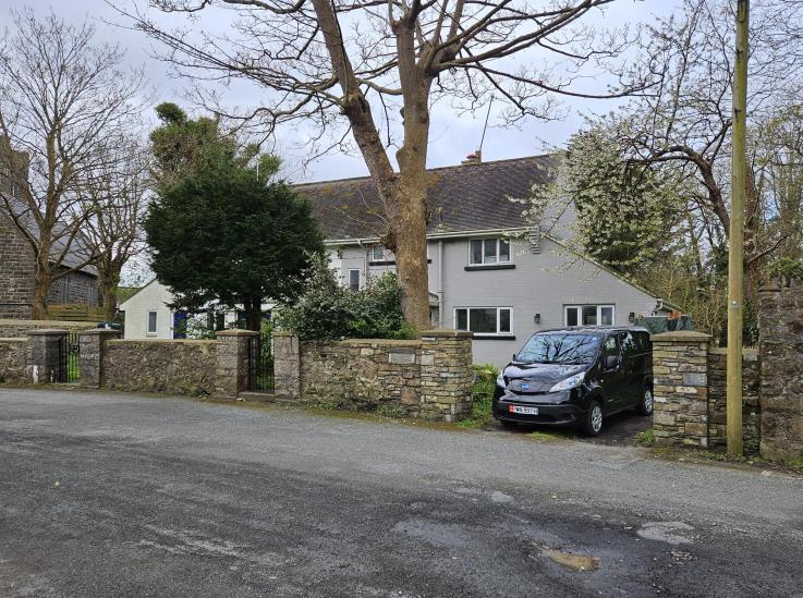 A street-level photograph showing a grey two-story detached house behind a stone wall and gate, with a black van parked in the driveway.