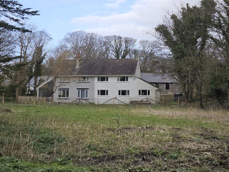 A photograph showing a white, two-story detached house situated in a rural setting with a grassy foreground and trees in the background.