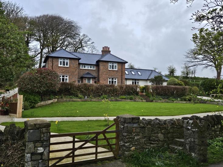 A photograph showing a large detached brick house with a slate roof set back on a grassy slope, viewed through a stone wall and wooden gate.