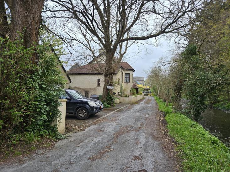 A street-level photograph showing a narrow rural road with a detached house on the left and a stream running alongside the road on the right.