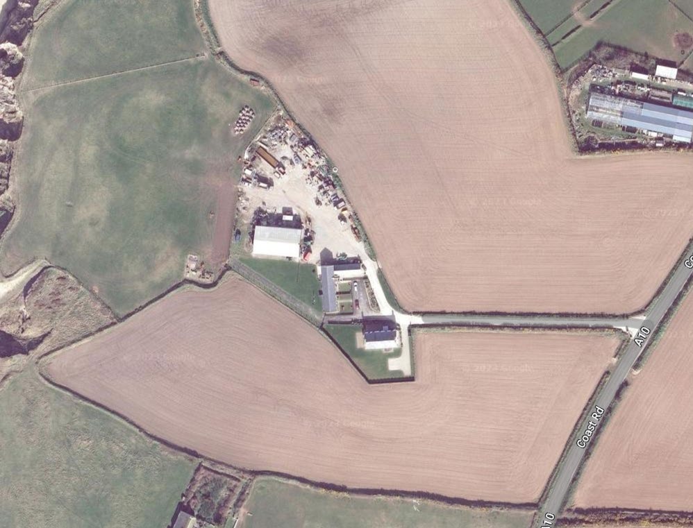 An aerial satellite view showing a rural site with buildings and storage areas adjacent to agricultural fields and the A10 Coast Road.