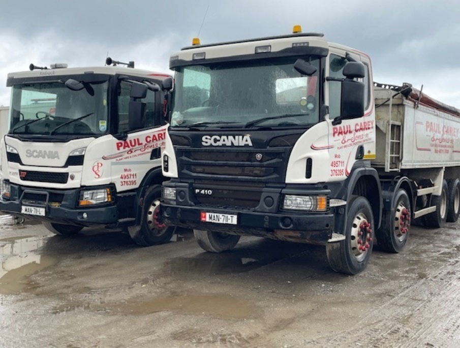 A photograph showing two white Scania trucks parked side-by-side on a muddy surface.
