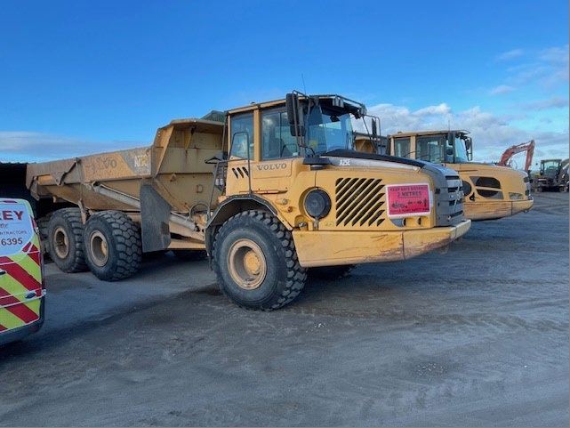 A photograph showing large yellow construction vehicles, including a Volvo articulated dump truck, parked in a gravel yard.