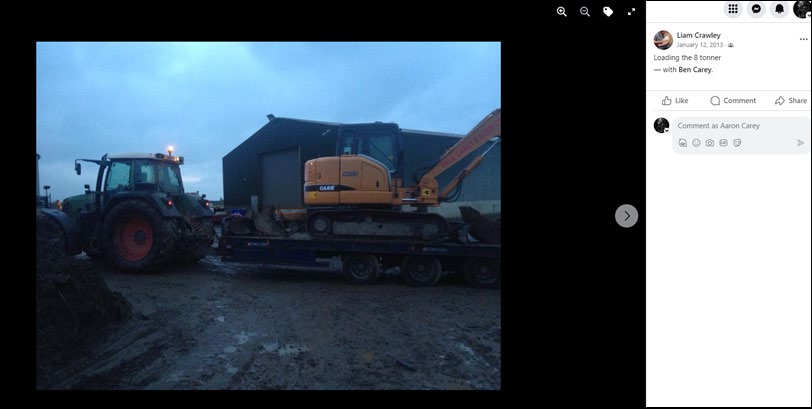 A photograph showing a tractor towing a trailer carrying an excavator, parked in front of a large industrial shed or workshop.