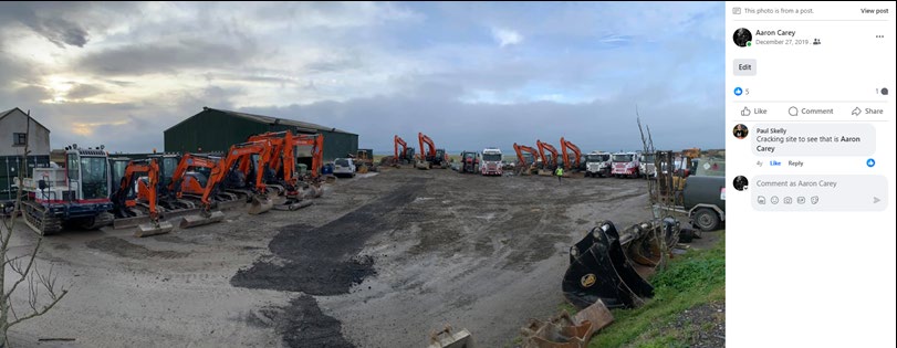 A panoramic photograph of an industrial yard or plant hire site featuring numerous orange excavators and trucks parked on a gravel surface in front of a large green warehouse shed.