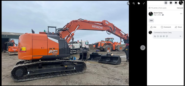 A photograph showing a large orange Hitachi excavator parked on a gravel yard, with other construction vehicles and a building visible in the background.