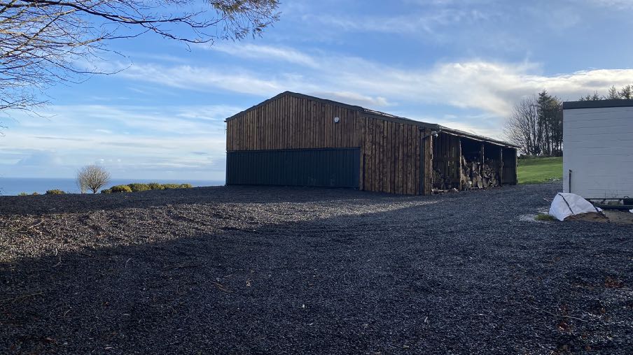 A photograph showing a large wooden outbuilding or barn situated on a gravel driveway with a view of the sea in the background.