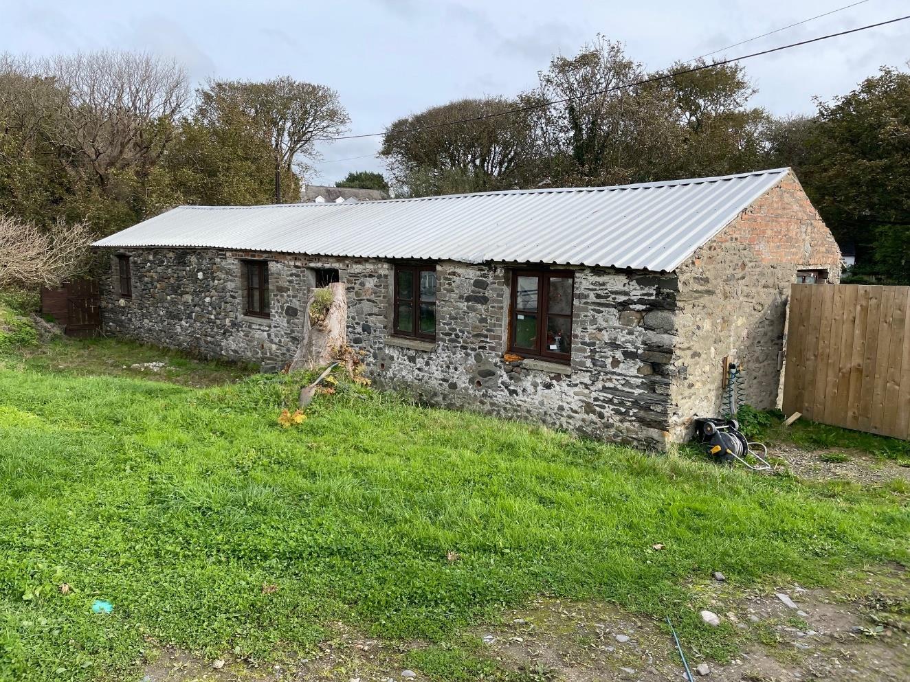 A photograph of a long, single-story stone building with a corrugated metal roof, situated in a grassy rural area with a wooden fence on the right.