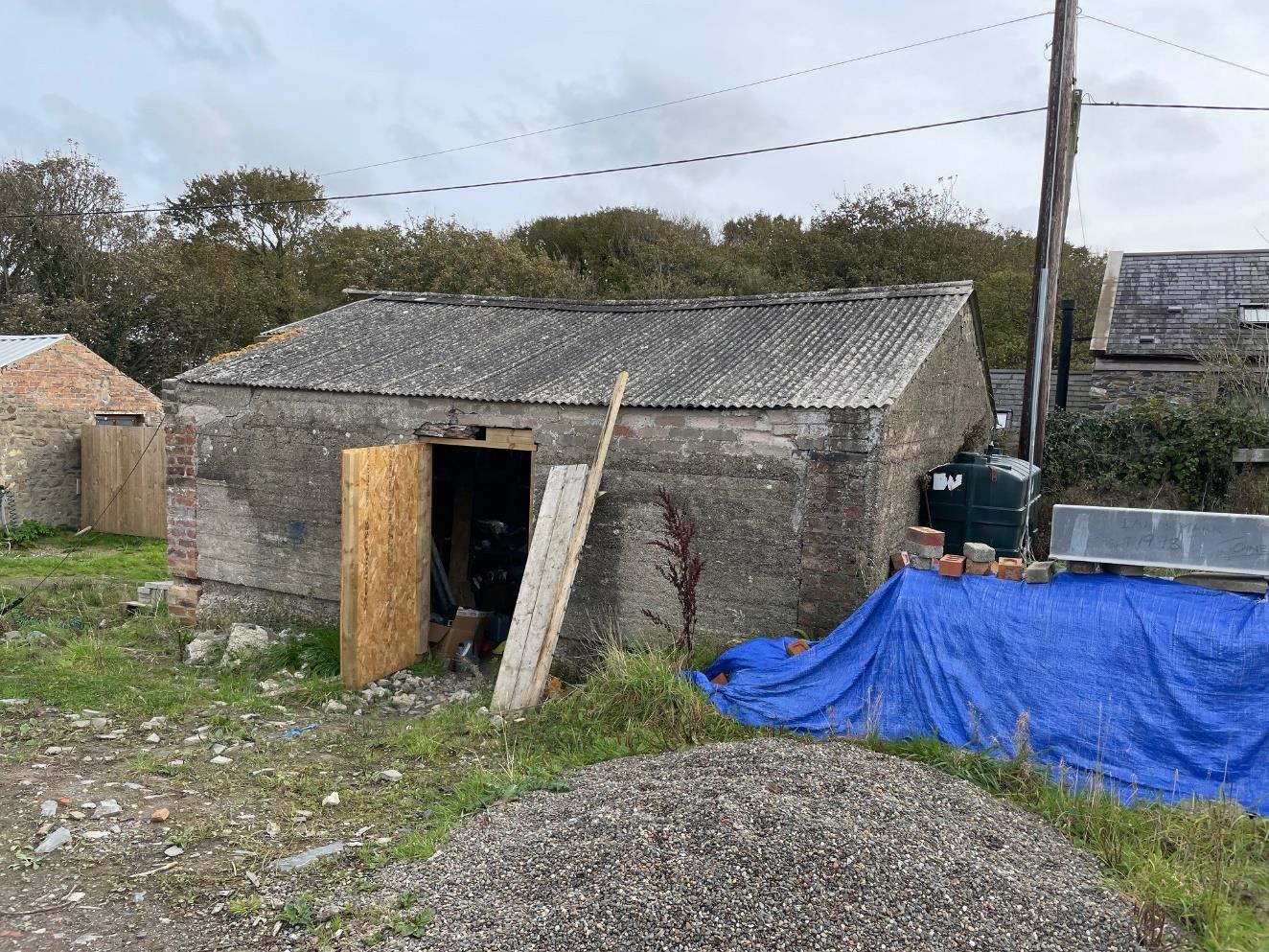 A photograph of a dilapidated stone outbuilding with a corrugated roof, partially boarded up and surrounded by construction materials like gravel and a blue tarp.