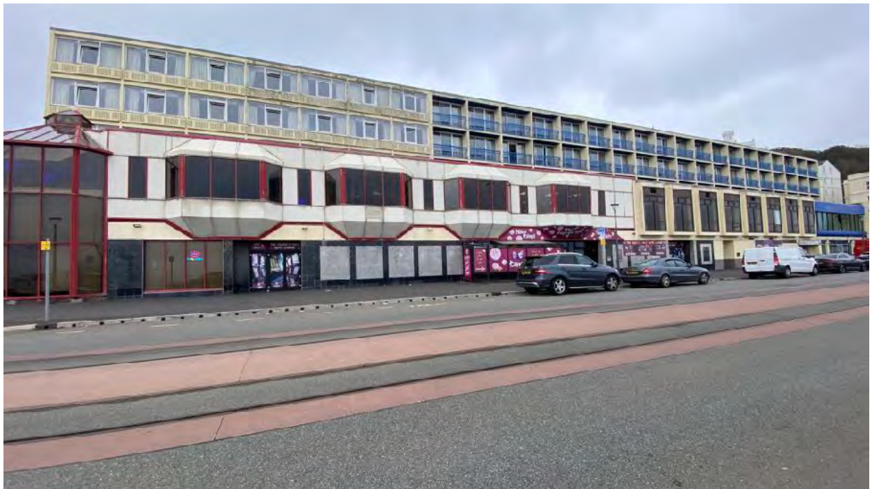A street-level photograph showing the exterior of a large, multi-story commercial building or hotel with balconies and ground-floor units.
