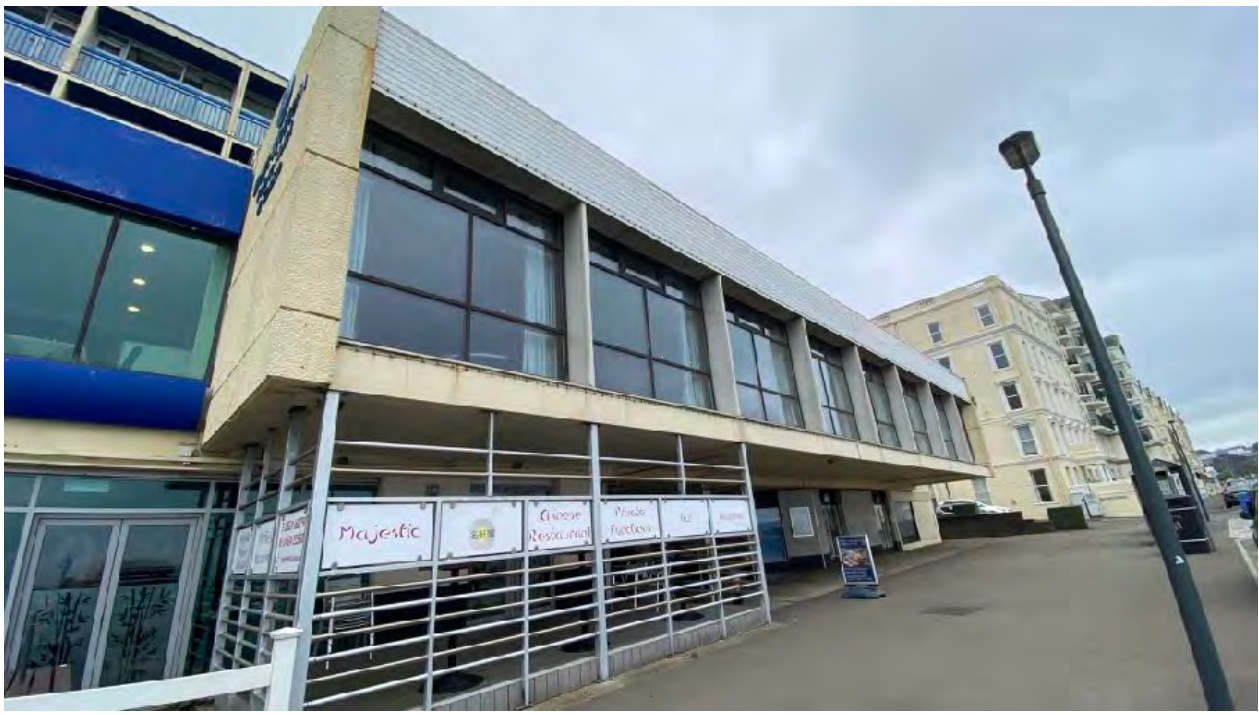 A street-level photograph showing a commercial building with a restaurant on the ground floor and large windows on the upper level, situated on a paved street next to other buildings.