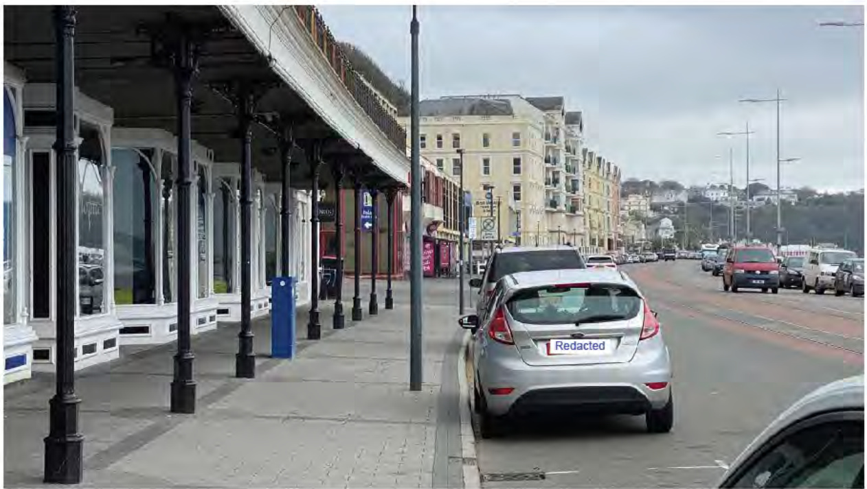 A street-level photograph of the Douglas promenade featuring a covered arcade with black pillars on the left and a road with traffic on the right.
