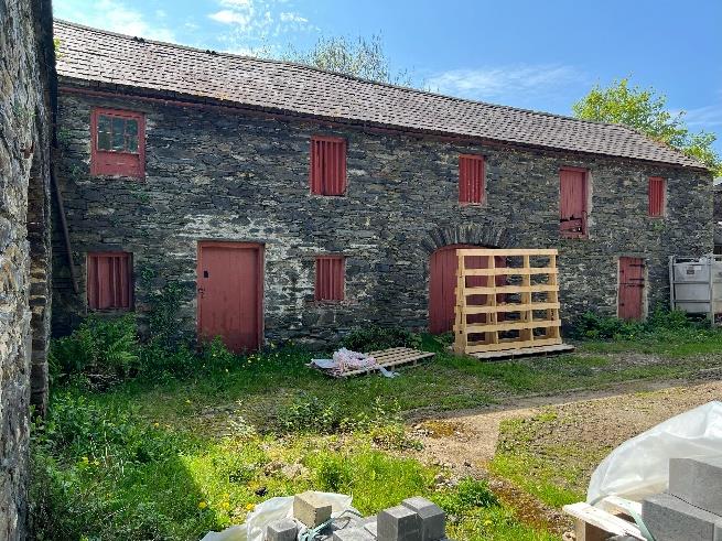 A photograph showing the exterior of a large, two-story stone barn with red wooden doors and shutters, set in a rural environment with construction materials visible in the foreground.