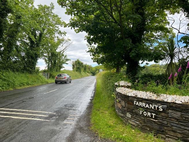 A photograph of a rural road scene featuring a stone boundary wall with the sign 'FARRANTS FORT' and a car driving past.