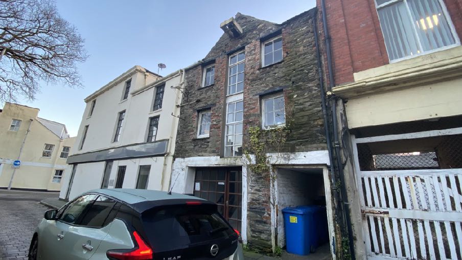 A street-level photograph showing a tall, narrow stone building with a gable end, situated between a white building and a red brick structure, with a car parked in the foreground.