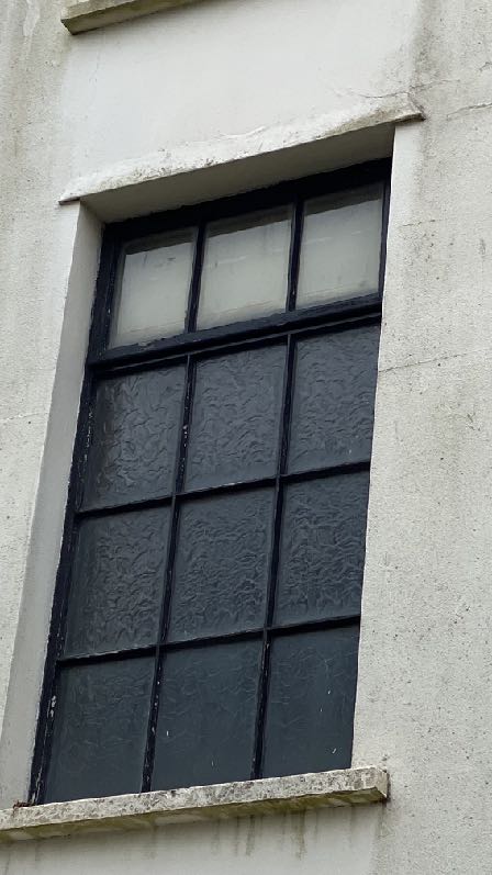 A close-up photograph showing a window with a black frame and textured glass set into a white rendered wall with a stone lintel and sill.