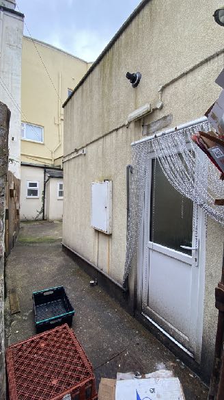A photograph showing the narrow side passage and rear wall of a building, featuring a white door with a beaded curtain and utility boxes.