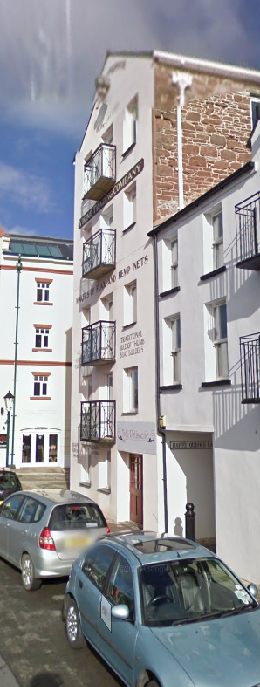 A street-level photograph showing a row of buildings, including a white building with balconies and a sign, alongside a building with exposed stone upper walls. Cars are parked on the street in the foreground.