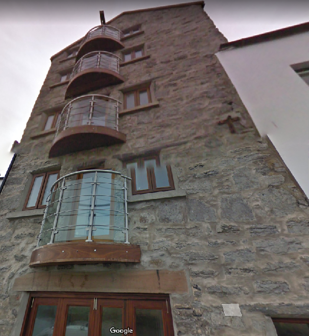 A low-angle photograph of a tall, multi-story stone building featuring curved balconies and wooden window frames, likely a converted barn or substantial house.
